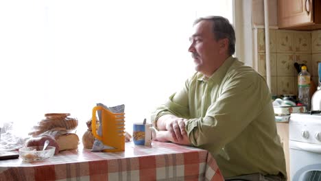 An-elderly-man-with-a-mustache-holds-a-cup-of-hot-drink-and-drinks.-He-sits-near-the-window-and-has-breakfast-at-home