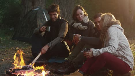 Group-of-people-sit-in-a-forest-next-to-a-campfire-with-warm-drinks-from-thermos-and-use-tablet.