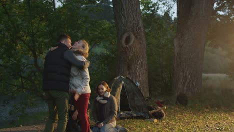 Man-arrives-to-the-campside-where-his-friends-are-sitting-next-to-campfire-and-a-girl-meets-him-with-a-hug.