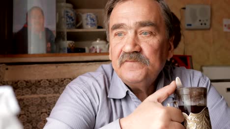 An-elderly-man-with-a-mustache-holds-a-cup-of-hot-drink-and-drinks.-He-sits-near-the-window-and-has-breakfast-at-home