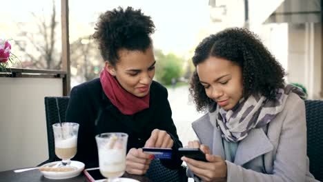 Two-attractive-mixed-race-women-having-online-shopping-with-credit-card-and-smartphone-while-talking-and-drinking-coffee-in-street-cafe