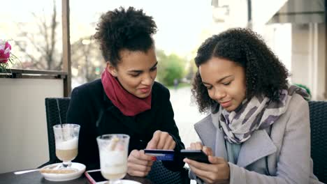 Zwei-attraktive-gemischte-Rasse-Frauen-mit-Online-Shopping-mit-Kreditkarte-und-Smartphone-beim-Sprechen-und-Kaffee-trinken-in-Straßencafé