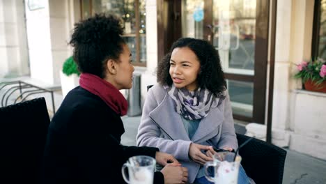 Dos-atractivas-mujeres-de-raza-mixta-hablando-y-bebiendo-café-en-el-café-de-la-calle.-Los-amigos-se-divierten-después-de-visitar-la-venta-de-centros-comerciales