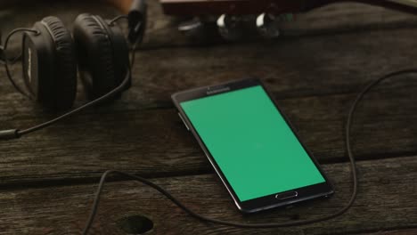 Android-Handy-mit-Greenscreen-im-Portrait-Modus-Handauflegen-Holztisch-neben-Gitarre-und-Kopfhörer.-Kausale-Lifestyle