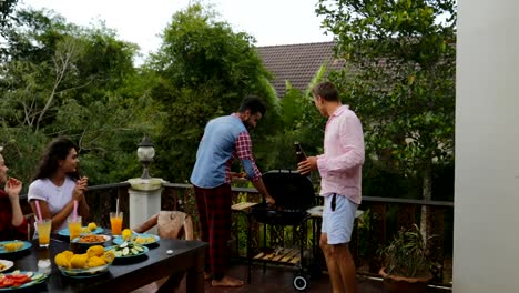 Two-Man-Taking-Food-From-Barbecue-People-Sitting-At-Table-Young-Friends-Group-Gathering-On-Summer-Terrace-Having-Party