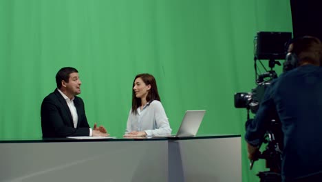 Backstage-footage-of-male-and-female-broadcasters-who-sit-at-a-table-and-talk-on-a-mock-up-green-screen-in-the-background.