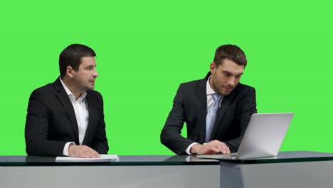 Two-media-broadcasters-are-sitting-at-a-table-with-a-laptop-and-talking-on-a-mock-up-green-screen-in-the-background.