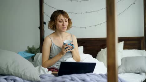 Attractive-young-woman-having-online-video-chat-with-friends-using-laptop-camera-while-sitting-on-bed-at-home