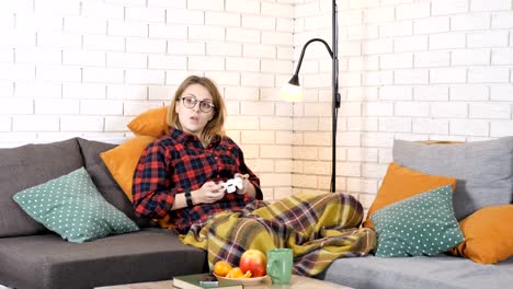 Cute-caucasian-girl-sitting-on-the-couch-under-the-blanket-and-playing-the-console-game,-yawns-50-fps