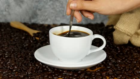 4k-Nahaufnahme-weibliche-Hand-rühren-Cup-schwarz-Kaffee---weibliche-Hand-mit-Löffel-um-zu-rühren,-frischem-Boden-schwarz-weiße-Kaffeetasse-Untertasse-neben-Kaffee-in-Bohnen,-Kaffee-Zeit