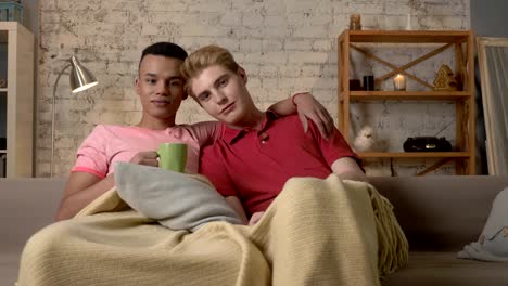 Two-gay-guys-sit-on-the-couch-and-watch-TV,-drink-hot-tea,-use-the-remote-control.-LGBT-lovers,-a-multinational-couple,-a-happy-gay-family,-a-home-cosiness-concept.-Look-at-the-camera-60-fps