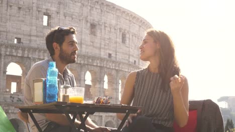 Glückliches-junges-Paar-Touristen-entspannende-chatten-Spaß,-sitzen-im-Restaurant-vor-dem-Kolosseum-in-Rom-bei-Sonnenuntergang-mit-Kaffee-beträufeln-und-Einkaufstaschen