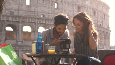 Jóvenes-felices-pareja-turistas-con-smartphone-sentado-en-bar-restaurante-frente-Coliseo-de-Roma-al-atardecer-con-bolsas-de-café-sonriendo-con-divertidos-mensajes-de-texto-de-navegación-y-compartir-fotografías