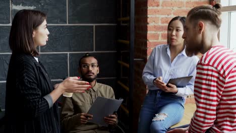 Group-of-multiracial-colleagues-disscussing-startup-project-while-standing-near-window-in-office-with-coffee-and-papers