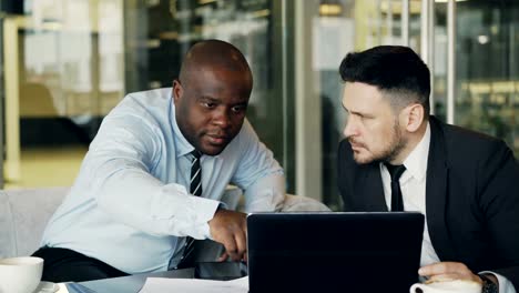 Two-business-colleagues-looking-at-laptop-computer-and-discussing-their-projects-in-modern-office-with-glass-walls.-Bearded-businessman-and-his-partner-sitting-at-table-and-talking