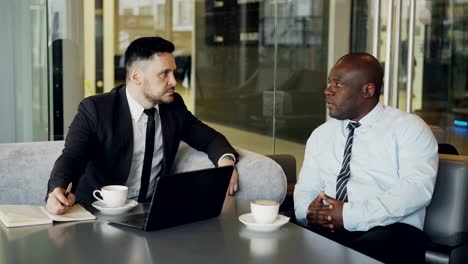 Two-confident-businessmen-discussing-about-partnership-during-meeting-in-modern-glassy-cafe