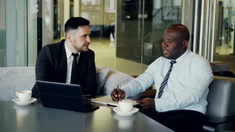 Two-cheerful-businessmen-discussing-project-in-modern-cafe.-African-American-entrepreneur-writing-in-notebook-and-his-caucasian-partner-in-business-suit-pointing-at-laptop-computer