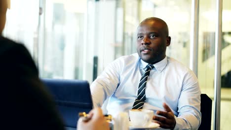 Hombre-de-negocios-afroamericano-gesticulando-y-explicando-detalles-de-su-arranque-inversor-en-desgaste-formal-en-café-vidrioso-durante-la-hora-del-almuerzo