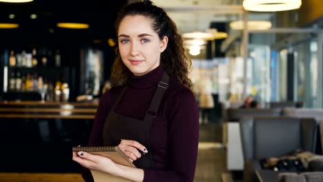 Portrait-of-cheerful-and-beautiful-young-waitress-standing,-keeping-menu,-and-smiling-sincerely-in-slylish-cafe.