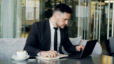 Smart-Caucasian-businessman-using-laptop-computer-and-writing-down-information-in-his-notepad-in-glassy-cafe-having-coffee-cup-on-his-table.