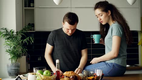 Happy-young-couple-cooking-ant-talking-in-the-kitchen-at-home.-Attractive-man-feeding-his-girlfriend-while-cutting-vegetables-for-breakfast-in-the-morning