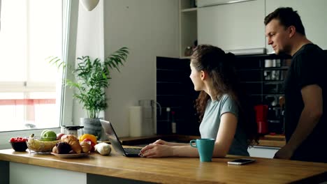 Junge-lockig-ziemlich-positiven-kaukasischen-Frau-sitzt-am-Tisch-und-haben-Videoanruf-auf-Laptop-Computer,-sie-sitzen-in-der-Küche-in-der-Nähe-von-Fenster,-ihr-Freund-kommt-und-sie-Skype-in-positiver-Weise-zusammen