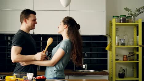 Pareja-feliz-divertirse-en-la-cocina-de-esgrima-con-grandes-cucharas-y-abrazándose-unos-a-otros-mientras-se-cocina-el-desayuno-en-casa