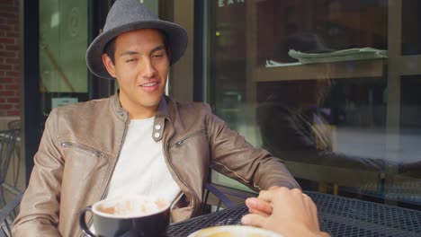 Young-man-sitting-outside-a-coffee-shop-smiles-and-holds-his-girlfriend's-hand,-portrait