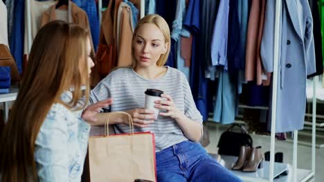 Young-woman-sitting-in-clothes-shop,-holding-coffee-and-talking-to-her-female-friend.-Ladies-having-rest-after-going-shopping.