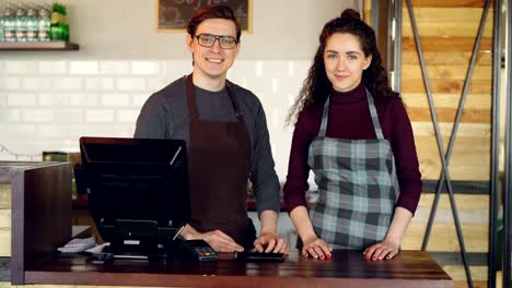 Portrait-of-two-partners-small-business-owners-standing-at-cashier's-desk-in-coffee-house-and-smiling.-Successful-business,-happy-people-and-food-service-concept.