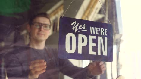 Attractive-male-waiter-in-apron-is-changing-doorplate-from-"yes-we-are-open"-to-"sorry-we-are-closed".-End-of-work-day,-employee-and-business-concept.