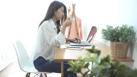 Beautiful-young-smiling-woman-working-on-laptop-while-enjoying-drinking-warm-coffee-sitting-in-a-living-room-at-home.-Enjoying-time-at-home.-Asian-business-woman-working-in-her-home-office.