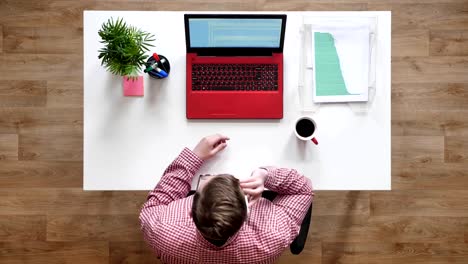 Young-man-in-glasses-typing-on-laptop-and-picking-up-phone,-topshot,-sitting-behind-desk-with-coffee-and-documents