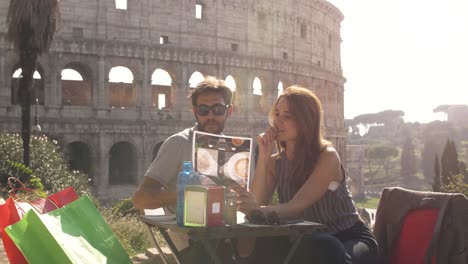 Happy-young-couple-tourists-reads-menu-choosing-food-and-drinks-sitting-at-bar-restaurant-in-front-of-colosseum-in-rome-at-sunset-ready-to-order