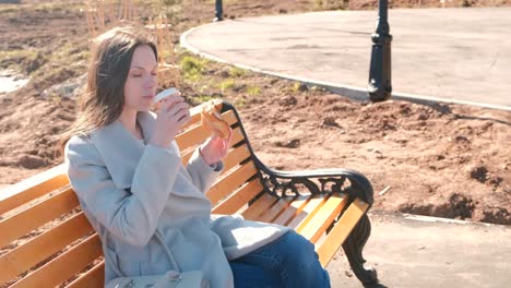 Young-brunette-woman-in-coat-sits-on-bench-in-city-park,-eats-bun-with-sugar-and-drinks-coffee.-Springtime.