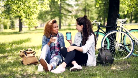 Alegre-jovencita-es-hablando-con-su-amiga-afroamericana-y-beber-café-para-llevar-en-el-parque-en-césped-verde-agradable.-Las-niñas-están-charlando-y-disfrutando-de-la-bebida.