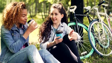 Alumnas-están-charlando-y-tomando-café-para-llevar-en-Parque-descanso-sobre-la-hierba-después-de-montar-en-bici.-Chicas-atractivas-están-sonriendo-y-riendo-disfrutando-de-bebidas.