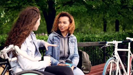 Young-women-tourists-are-having-conversation-sitting-on-bench-in-park-and-holding-takeaway-coffee-with-bikes-standing-nearby.-Tourism,-people-and-communication-concept.