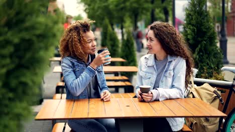 Fröhliche-Junge-Frauen-Beste-Freunde-lachen-und-reden-beim-Sitzen-am-Tisch-im-Café-im-freien-und-Kaffee-zu-trinken,-sind-Mädchen-Geselligkeit-und-Spaß-Neuigkeiten-diskutieren.