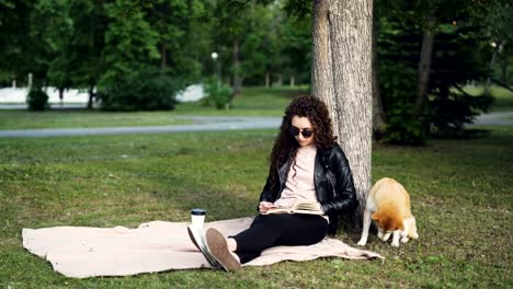 Relaxed-girl-is-reading-book-and-drinking-takeaway-coffee-sitting-on-lawn-in-park-while-her-cute-dog-is-smelling-ground-and-air-sitting-under-tree.-Leisure-and-pets-concept.