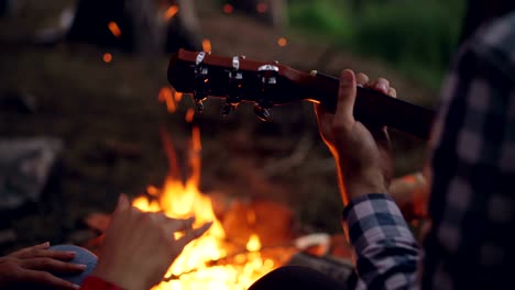 Toma-de-primer-plano-de-manos-hombres-tocando-la-guitarra-y-la-mujer-las-manos-en-el-baile-con-ardiente-fogata-en-fondo.-Camping,-concepto-de-música-y-gente.