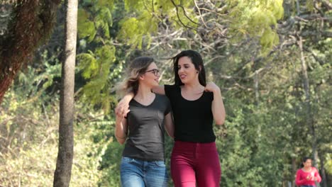 Two-girlfriends-together-walking-at-the-park-embracing-each-other-laughing