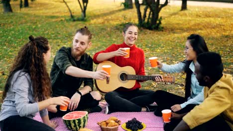 Cheerful-young-people-multi-ethnic-group-are-clinking-glasses-with-drinks-then-drinking-sitting-on-blanket-on-grass-during-autumn-picnic-with-guitar-and-food.