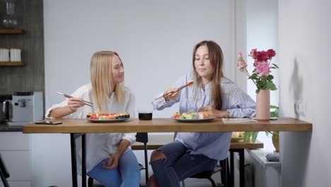 Two-young-women-talking-and-eating-sushi-at-home,-best-friends-spending-time-together