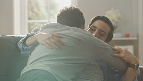 Süße-männliche-Gay-paar-voll-um-auf-dem-Sofa-zu-Hause.-Freund-läuft-und-springt-in-die-Hände-seines-Partners.-Sie-umarmen-sich.-Sie-sind-glücklich-und-lachend.-Sie-sind-lässig-gekleidet-und-Zimmer-ist-Modern-eingerichtet.