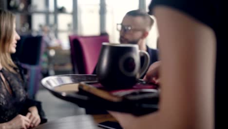 El-camarero-lleva-un-vaso-de-cappuccino-grande-para-la-joven-pareja-en-la-bandeja,-que-realiza-un-aperitivo-en-un-restaurante,-un-hombre-y-una-mujer-hablan-sobre-el-pasado-fin-de-semana