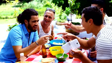 Smiling-man-showing-his-mobile-phone-to-his-friends-while-having-meal-outdoors