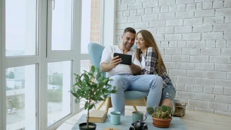 Young-attractive-couple-having-online-video-chat-using-tablet-computer-sitting-at-balcony-in-modern-loft-apartment