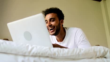 Young-Man-Using-Laptop-Computer-Lying-On-Bed-Happy-Smiling-Hispanic-Guy-Chatting-Online-In-Bedroom-Morning