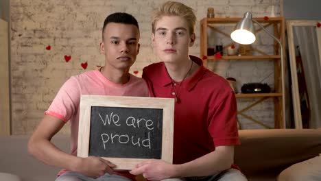 A-sad-international-gay-couple-is-sitting-on-the-couch-and-holding-a-sign.-We-are-proud.-Look-at-the-camera.-Home-comfort-on-the-background.-60-fps